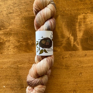 May include: A skein of hand-dyed yarn in shades of brown, beige, and cream. The yarn is wrapped around a white label with a brown bird's nest and the text "MADE BY RABBIT" and "hand dyed in the foothills".
