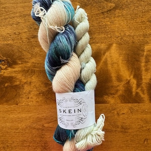 May include: Two skeins of yarn, one is a light blue, teal, and white variegated yarn, the other is a cream colored yarn. The yarn is wrapped around a label that reads "Australia SKEIN Artisan Yarn and Fibre Since 2010."