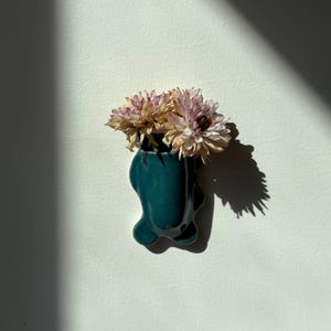 May include: A dark teal ceramic wall-mounted vase with an organic, asymmetrical shape. The vase holds a small bouquet of dried, pale pink and cream-colored flowers with textured, layered petals, casting a distinct shadow on the wall.