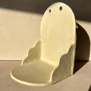 May include: A handmade, off-white ceramic wall shelf with a rounded top and a scalloped edge. The shelf has two small holes near the top for hanging. The design is minimalist and modern, suitable for displaying small items.