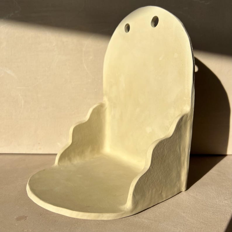 May include: A handmade, off-white ceramic wall shelf with a rounded top and a scalloped edge. The shelf has two small holes near the top for hanging. The design is minimalist and modern, suitable for displaying small items.