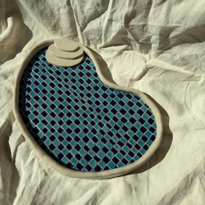 May include: A kidney-shaped ceramic tray with a glossy finish, featuring a grid pattern of alternating dark blue and turquoise square tiles. The tray has a raised, off-white border and a small, three-tiered, off-white staircase element.