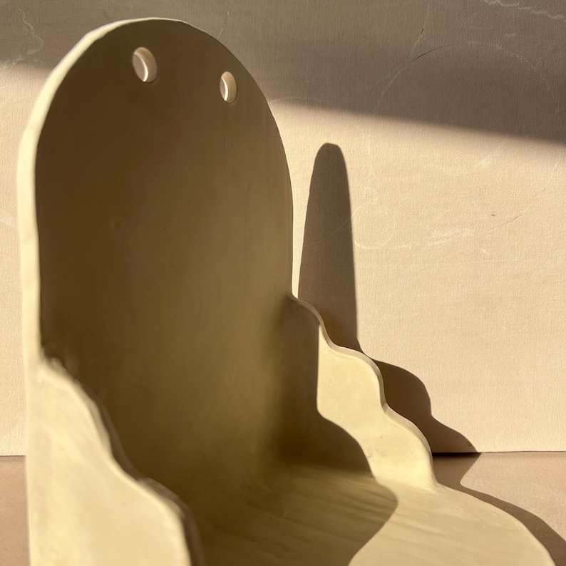 May include: A cream-colored, ceramic wall shelf with a unique, wavy design. The shelf has a curved back with two small, oval cutouts near the top. The shelf's edges have a soft, undulating shape, and it casts a long shadow.
