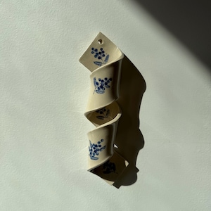 May include: A cream-colored ceramic wall vase with a spiraling, ribbon-like structure. The surface is decorated with small, dark blue floral sprig patterns. A single circular hole is visible at the top for mounting the piece to a wall.