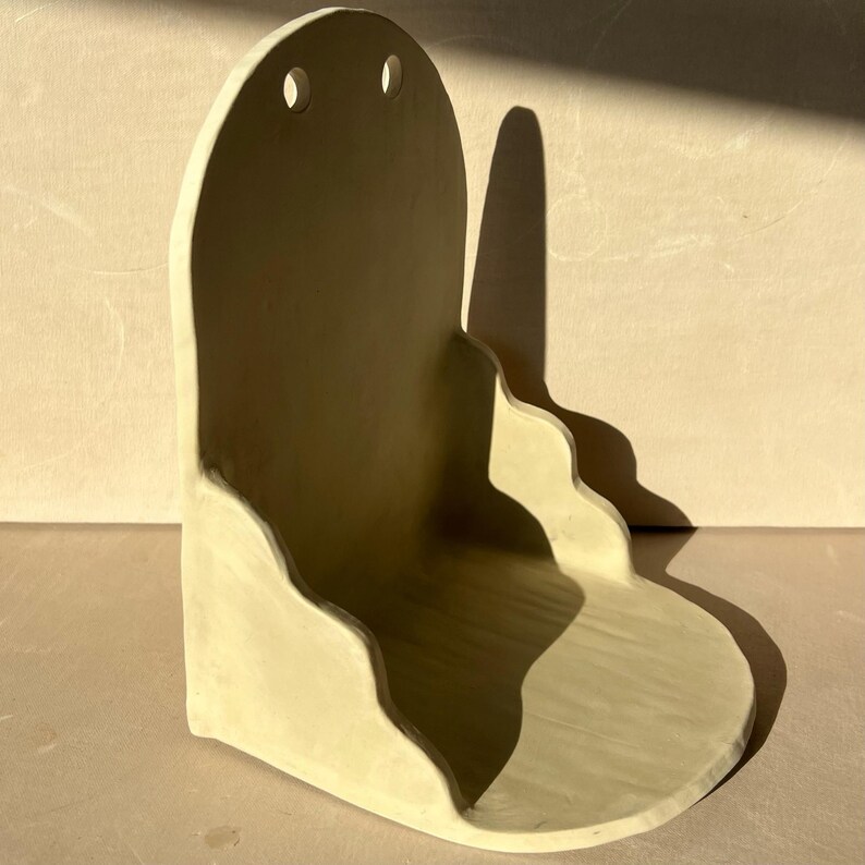 May include: A beige ceramic wall shelf with a curved design. The shelf has a rounded top with two small holes and a wavy bottom edge. The shelf is made of a light-colored clay and has a matte finish. The shelf is designed to be mounted on a wall.