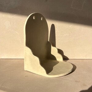 May include: A light beige ceramic wall shelf with a rounded top and two small holes. The shelf has a wavy design along the bottom and a small, rounded base. The shelf is designed to be mounted on a wall and is likely used for decorative items.