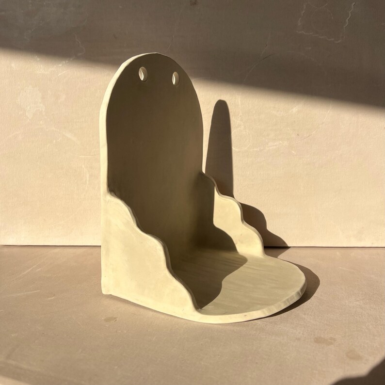 May include: A light beige ceramic wall shelf with a rounded top and two small holes. The shelf has a wavy design along the bottom and a small, rounded base. The shelf is designed to be mounted on a wall and is likely used for decorative items.