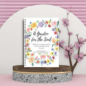 May include: A spiral-bound journal titled "A Garden for the Soul" with a floral wreath design. The journal is displayed on a wooden and stone pedestal, with a pink and white backdrop and a branch with pink flowers.