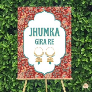 May include: A red and white sign with a paisley pattern and the text "Jhumka Gira Re" in teal blue. Two gold jhumka earrings with teal blue accents are pictured below the text.