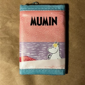 May include: A pink and blue wallet with a cartoon image of Moomin on the front. The wallet has the word "Moomin" printed in black letters.