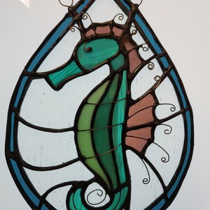 May include: A stained glass window hanging with a teal blue, green, and pink seahorse design.