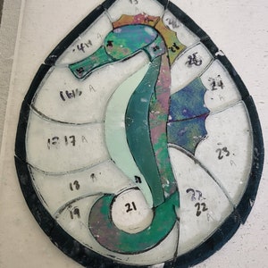 May include: A stained glass seahorse design with numbered pieces of glass. The seahorse is green, blue, and purple with a yellow and orange mane.