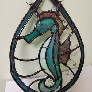 May include: A stained glass window hanging features a teal blue and green seahorse with a brown outline. The seahorse is surrounded by a teardrop-shaped frame with a blue background.