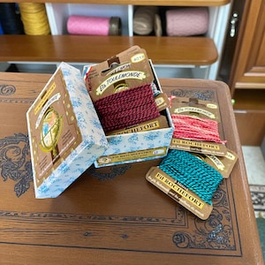 May include: A box with blue floral patterns holds spools of red, pink, and teal twine. The box is labelled "Fil Rochefort" and "Els Toulemonde". The twine spools are displayed on a wooden surface.