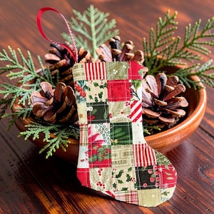 May include: A small quilted Christmas stocking ornament featuring a patchwork design of various holiday-themed fabrics in red, green, and cream. The fabric squares display patterns of holly, poinsettias, and text reading "ON E" and "HALLS".