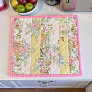 May include: A quilted placemat featuring a floral design in pink, yellow, and white. The placemat has a pink polka dot border and is divided into vertical sections. The floral design includes pink, blue, and green flowers.