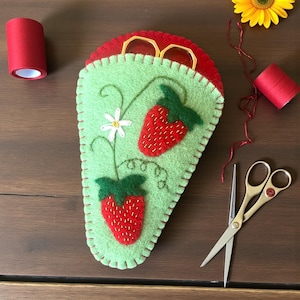 Puede incluir: Un kit de costura artesanal con temática de fresas de fieltro. El kit presenta una funda verde claro con fresas bordadas y una flor blanca. También hay tijeras doradas, carretes de hilo rojo y una flor amarilla.
