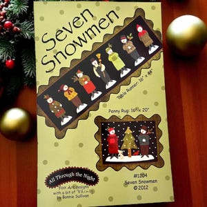 May include: A craft pattern titled "Seven Snowmen" featuring folk art designs. The cover displays a table runner design (40.6cm x 111.8cm) and a penny rug design (40.6cm x 50.8cm). The design includes seven snowman figures and a winter scene.