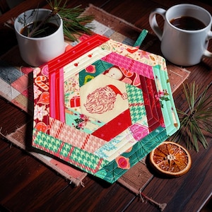 May include: A hexagonal quilted potholder featuring a central cream-coloured fabric panel with a red line-art illustration of Santa Claus. The surrounding border consists of various fabric strips in shades of red, green, and cream with holiday-themed patterns including candy canes, holly, and houndstooth.