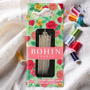 May include: A rectangular packet of Bohin France sewing needles featuring a green background with red and yellow rose illustrations. The packet displays the text BOHIN FRANCE and COUTURE COURANTE SHARPS FOR SEWING BIG EYES. Several small spools of thread are nearby.