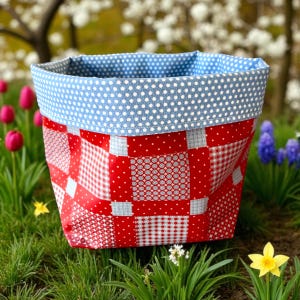 May include: A fabric storage basket with a patchwork design in red, white, and blue. The basket has a blue polka dot lining and is set in a garden with flowers and grass.