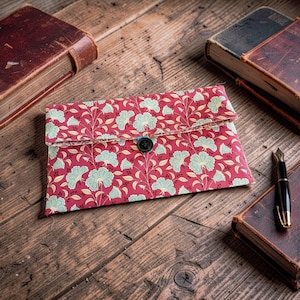 May include: A rectangular fabric envelope pouch featuring a repeating pattern of light green floral motifs on a deep red background. The pouch has a fold-over flap secured by a single black button centred on the front edge of the closure.