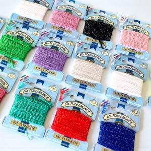 May include: A selection of Fil Tonkin thread spools in a variety of colours, such as pink, green, purple, and red. Each spool is labelled with the brand name and a product number. The threads are made of 100% polyester.