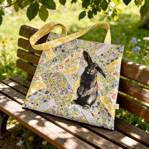 May include: A square tote bag featuring a patchwork design of various floral fabrics in shades of yellow, pink, blue, and green. A black rabbit illustration is centred on the front panel. The bag has two yellow floral fabric shoulder straps.