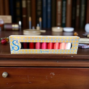 May include: A box of embroidery thread in shades of red, pink, and white. The box is yellow with a geometric pattern and a cutout to display the thread. The brand name "S" is visible on the box. The box is on a wooden surface.