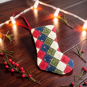 May include: A quilted fabric stocking ornament featuring a patchwork pattern of red, navy blue, and green squares with a small cherry print. The stocking has a red ribbon loop at the top for hanging and is stitched with a grid-like quilted texture.