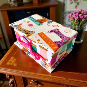 May include: A rectangular sewing box with a white background and a colourful pattern of sewing machines, spools of thread, and other sewing notions. The box is tied with a bright pink ribbon. The box is on a wooden table.