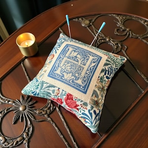 May include: A decorative pin cushion with a floral pattern and a central square displaying a blue letter "H" design. Two acupuncture needles are inserted. A small gold candle holder with a lit candle is in the background.