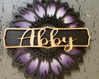 Op de afbeelding: Houten zonnebloem wanddecoratie met de naam "Abby" in sierlijke letters. De zonnebloem heeft een zwarte omtrek met paarse en witte bloemblaadjes. De naam staat op een zwart bord met een lichte houten rand.