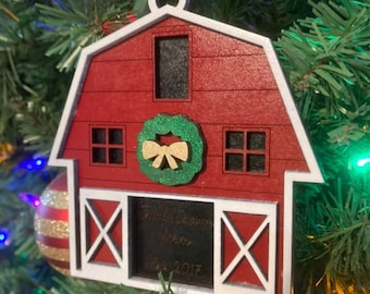 May include: A red barn-shaped Christmas ornament with white trim, a green wreath, and a gold bow. The ornament has a black area with engraved text and the year 2017. It hangs on a Christmas tree.