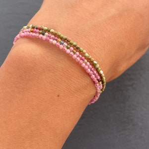 Peut inclure: Un bracelet superposé avec des perles roses, vertes et dorées. Le bracelet est composé de petites perles rondes et est porté au poignet.