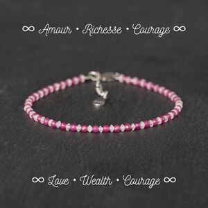 May include: A delicate bracelet made with pink and white beads. The bracelet has a silver clasp and the text "∞ Love • Wealth • Courage ∞" is printed below the bracelet.