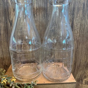 May include: Two clear glass milk bottles with a vintage design. The bottles are cylindrical with a narrow neck and a wide base. The word "OFFER" is embossed on the lower part of the bottles. The bottles are displayed on a wooden box.