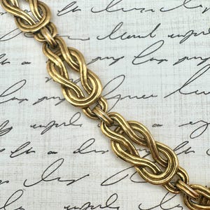 May include: A gold-toned metal chain necklace featuring a series of repeating infinity-style knot links. Each link is constructed from two intertwined loops of polished metal, creating a continuous, textured pattern throughout the entire length.