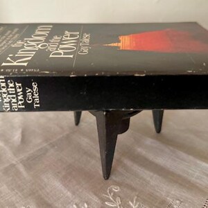 Puede incluir: Un libro negro de tapa dura titulado "The Kingdom and the Power" de Gay Talese. El libro est&aacute; sobre un soporte de metal negro con tres patas.
