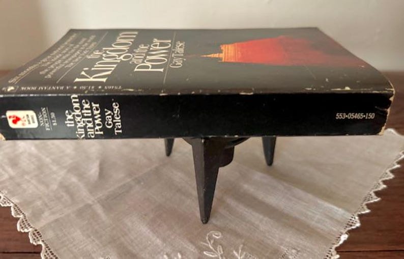 Puede incluir: Un libro negro de tapa dura titulado "The Kingdom and the Power" de Gay Talese. El libro est&aacute; sobre un soporte de metal negro con tres patas.