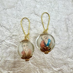 May include: A pair of drop earrings featuring clear spherical glass beads, each containing a miniature hand-painted portrait of an elderly person. The beads are accented with gold-toned metal caps at the base and suspended from gold-toned wire hooks.