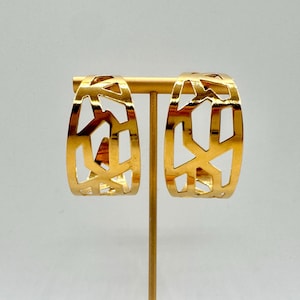 May include: A pair of gold-toned hoop earrings featuring an openwork geometric pattern with irregular cutouts. The earrings are displayed on a gold-colored T-shaped stand, showcasing a polished metallic finish and a wide, curved silhouette.