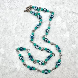 May include: A necklace featuring a series of round glass evil eye beads in shades of blue and white, interspersed with smaller solid green beads and silver-toned metal chain links. The piece is secured with a silver-toned metal toggle clasp.