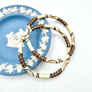 May include: Two ivory-colored bangles with bronze-colored metal accents and decorative detailing. The bangles are resting on a blue plate with white relief designs. The plate has a classical design with figures and floral patterns.