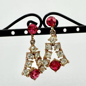 May include: A pair of gold-tone clip-on earrings featuring a triangular openwork design. Each earring is adorned with clear white rhinestones and accented with two round, vibrant pink crystals, one at the top and one at the base.