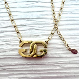 May include: A gold-toned necklace featuring a double "G" pendant. The pendant is the focal point, with a chain that extends to an adjustable length. The necklace is set against a textured white background.