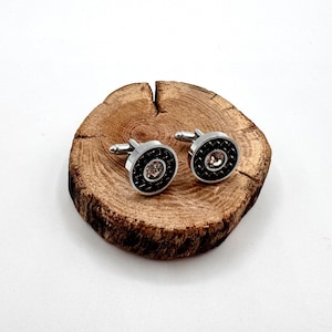 May include: A pair of silver cufflinks with a black, textured outer ring and a clear, round center stone. The cufflinks are displayed on a small, round, wooden surface with a natural wood grain.
