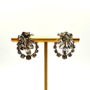 May include: A pair of vintage silver-toned clip-on earrings. Each earring features a circular design with a cluster of clear, faceted stones at the top and a ring of smaller stones around the bottom. The earrings are displayed on a gold-colored stand.