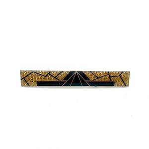 May include: A rectangular hair barrette with a geometric design. The barrette features a gold-colored base with small, evenly spaced embellishments. Black lines and a dark teal section create a contrasting geometric pattern. The barrette is set against a white background.