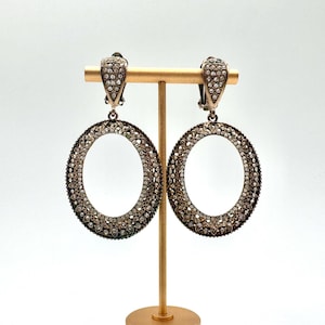 May include: A pair of oval-shaped clip-on earrings. The earrings are silver-toned and encrusted with small, clear crystals. They are displayed on a gold-colored earring stand against a white background. The earrings have a vintage or antique aesthetic.